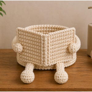 May include: A cream-colored, crocheted basket shaped like a book, with arms and legs. The basket is open at the top and has a book-like appearance, with the pages open. The arms and legs are also crocheted and add a whimsical touch to the design.