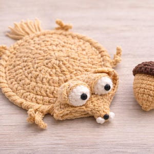 May include: Crocheted Scrat from Ice Age with a crocheted acorn. The Scrat design is tan with white eyes and black pupils. The acorn is tan with a brown cap. The items are on a light wood surface.