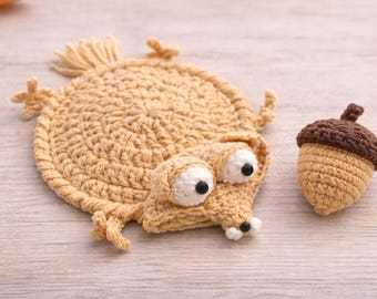 May include: Crocheted Scrat from Ice Age with a crocheted acorn. The Scrat design is tan with white eyes and black pupils. The acorn is tan with a brown cap. The items are on a light wood surface.