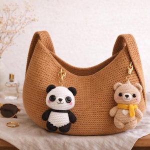 May include: A crocheted brown hobo bag with two attached keychains. One keychain is a panda bear, the other is a teddy bear. The panda is black and white, and the teddy bear is tan with a yellow scarf.