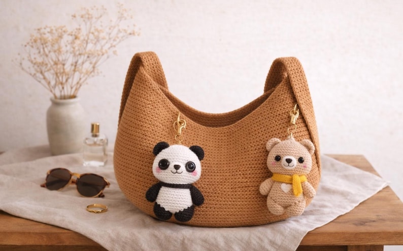 May include: A crocheted brown hobo bag with two attached keychains. One keychain is a panda bear, the other is a teddy bear. The panda is black and white, and the teddy bear is tan with a yellow scarf.