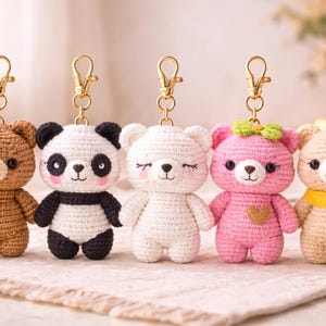May include: Five crochet bear keychains in various colors: brown, panda, white, pink, and tan. Each bear has a gold-colored clasp. The pink bear has a green bow and a heart. The tan bear has a yellow scarf.