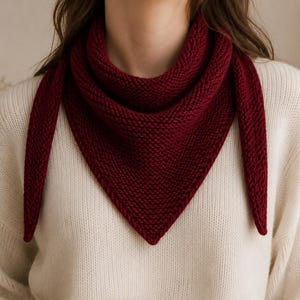 May include: A knitted burgundy scarf is draped around the neck, creating a triangular shape. The scarf is made of a thick, textured yarn and contrasts with the off-white sweater underneath. The scarf's ends hang down, adding a touch of elegance.