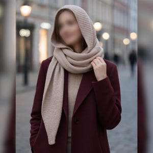 Puede incluir: Una persona con un abrigo burdeos, un suéter beige y una bufanda beige con capucha. La bufanda está envuelta alrededor del cuello y los hombros. El fondo está borroso, con farolas y edificios visibles.