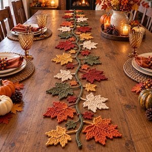 May include: A long, decorative table runner featuring a central crocheted vine adorned with various maple leaves in shades of red, orange, yellow, green, and cream. The runner is displayed on a wooden dining table set with plates, napkins, and glassware.