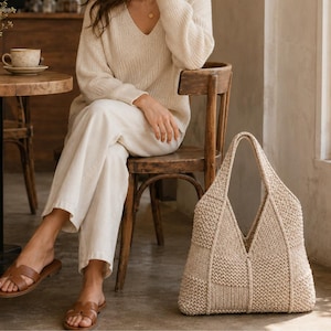 Könnte beinhalten: Eine beige Strick-Tragetasche mit Patchwork-Design und verschiedenen Strickmustern. Die Tasche hat eine strukturierte V-förmige Öffnung und zwei integrierte Griffe, gefertigt aus einem neutralfarbenen Garn mit texturierter Optik.