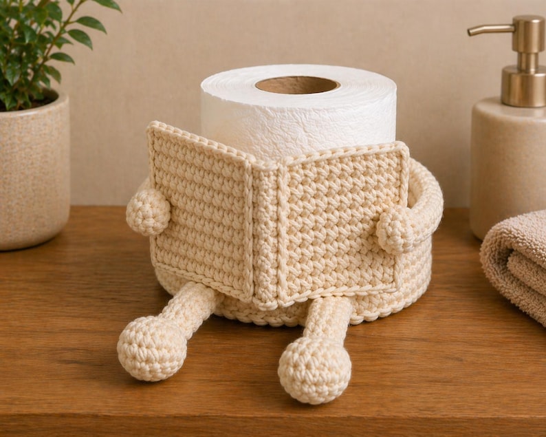 May include: A cream-colored, crocheted toilet paper holder shaped like a person reading a book. The holder has arms, legs, and a book-shaped cover, with a roll of white toilet paper inside. The holder is sitting on a wooden surface.