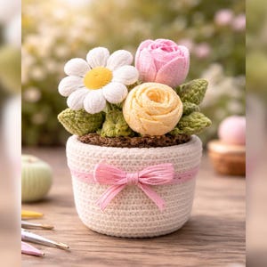 Może przedstawiać: Szydełkowa kompozycja kwiatowa w białej doniczce z różową kokardą. Kompozycja zawiera białą stokrotkę, różową różę i żółtą różę, z zielonymi liśćmi. Doniczka stoi na drewnianej powierzchni.