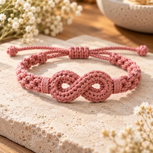May include: A dusty rose coloured bracelet crafted from woven cord featuring a central infinity symbol knot design. The bracelet includes an adjustable sliding knot closure with two small knotted ends, showcasing a detailed braided texture throughout.