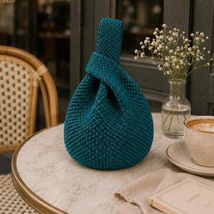 May include: A teal-colored, hand-knitted bag with a knotted handle sits on a marble-topped table. The bag is next to a cup of coffee, a small vase of white flowers, and a book. The bag's texture is visible, and the overall aesthetic is cozy.