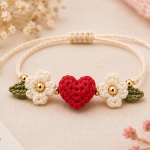 May include: A bracelet featuring a central red crocheted heart flanked by two white crocheted flowers and two small green crocheted leaves. The design includes small gold-toned beads and is held together by a light beige cord with adjustable knots.