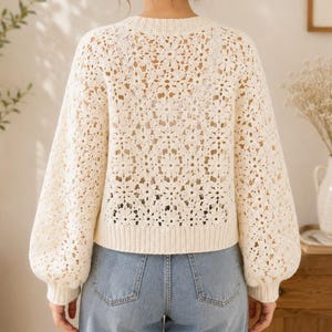 May include: Cream-colored crochet sweater with a floral pattern. The sweater has long, puffy sleeves and a ribbed hem. The model is wearing blue jeans. The sweater is a light, airy knit, perfect for layering.