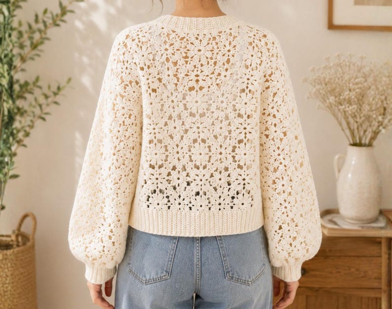 May include: Cream-colored crochet sweater with a floral pattern. The sweater has long, puffy sleeves and a ribbed hem. The model is wearing blue jeans. The sweater is a light, airy knit, perfect for layering.