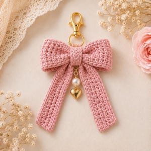 Puede incluir: Un llavero de lazo tejido a ganchillo en color rosa empolvado con un cierre de mosquetón dorado. Una pequeña cadena dorada cuelga del centro del lazo, adornada con una perla blanca artificial y un pequeño dije liso en forma de corazón.