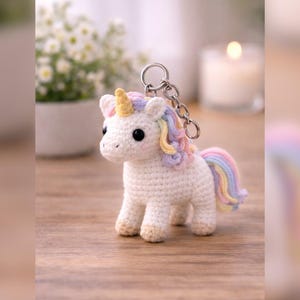 Könnte beinhalten: Ein weißer gehäkelter Einhorn-Schlüsselanhänger mit goldenem Horn, Regenbogenmähne und -schwanz sowie schwarzen Augen. Das Einhorn steht auf einer Holzoberfläche. Ein silberner Schlüsselanhänger ist oben am Einhorn befestigt.