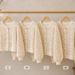 May include: Four cream-colored, open-weave knit cardigans hang on a wooden rod. Each cardigan has a round neckline, button closures, and long, puffy sleeves. Sizes S, M, L, and XL are indicated below each cardigan.