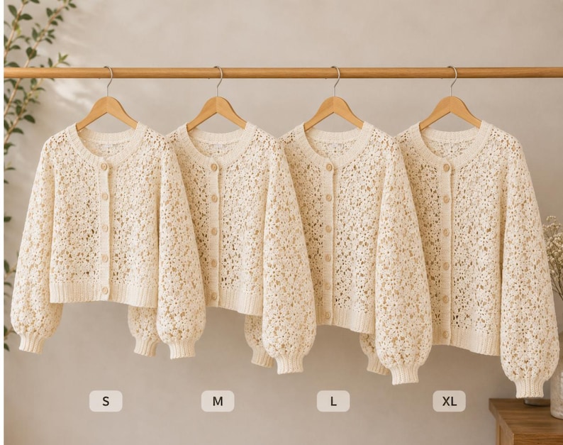 May include: Four cream-colored, open-weave knit cardigans hang on a wooden rod. Each cardigan has a round neckline, button closures, and long, puffy sleeves. Sizes S, M, L, and XL are indicated below each cardigan.