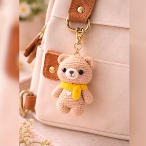 May include: A crocheted teddy bear keychain with a yellow scarf. The bear is light brown with black eyes and pink cheeks. It hangs from a gold-colored clasp attached to a cream-colored backpack.