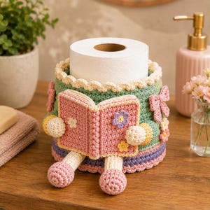 May include: A crochet toilet paper roll cover featuring a sage green base with a scalloped cream top edge. A pink crocheted book is attached to the front, held by two small cream-colored hands, with pink legs and feet extending from the bottom.
