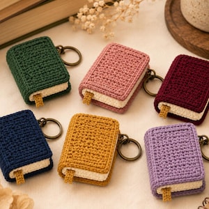 May include: Six miniature book-shaped keychains crafted from crochet yarn in dark green, dusty pink, deep burgundy, navy blue, mustard yellow, and light purple. Each keychain features a cream-colored page section and a small matching bookmark tab.
