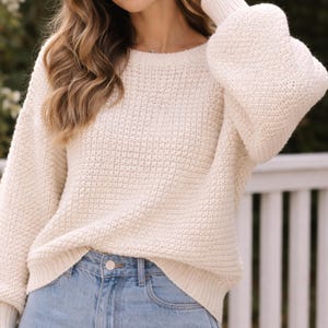 May include: Cream-colored knit sweater with a round neckline and long sleeves. The sweater has a textured knit pattern and is paired with blue denim jeans. The sleeves have a balloon shape at the upper arm and taper to ribbed cuffs.