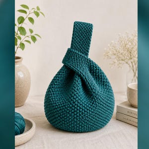 May include: A teal-colored, hand-knitted bag with a knotted handle. The bag has a rounded shape and a textured surface. The handle is wide and integrated into the bag's design. The background includes a vase with greenery and a ball of yarn.