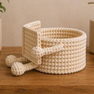 May include: A cream-colored, crocheted basket with a unique design. The basket features a round base and a rectangular handle, with two rounded feet. The basket is made of a thick, textured yarn and sits on a wooden surface.