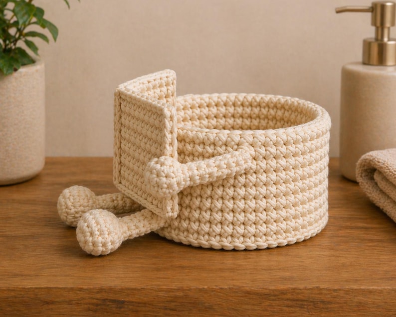 May include: A cream-colored, crocheted basket with a unique design. The basket features a round base and a rectangular handle, with two rounded feet. The basket is made of a thick, textured yarn and sits on a wooden surface.