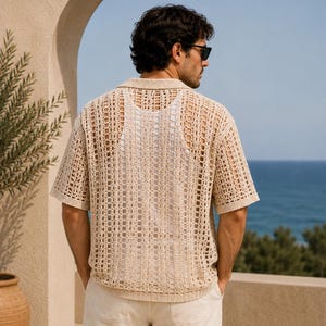 May include: A beige, open-weave polo shirt with short sleeves. The shirt has a collared neckline and a loose, breathable design. The person is wearing sunglasses and white pants. The background includes a view of the ocean and a beige archway.