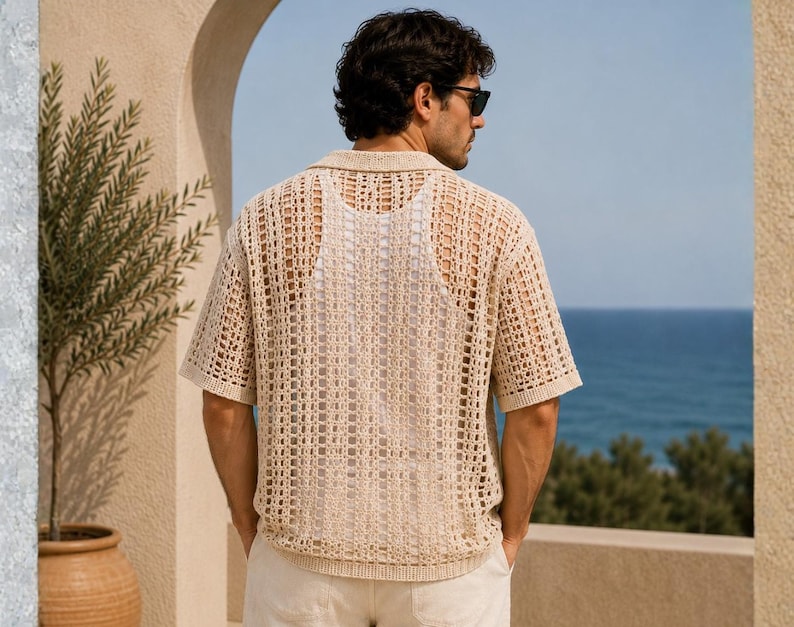 May include: A beige, open-weave polo shirt with short sleeves. The shirt has a collared neckline and a loose, breathable design. The person is wearing sunglasses and white pants. The background includes a view of the ocean and a beige archway.