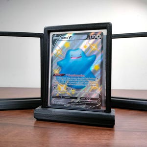 May include: Black display frame holding a Pokémon trading card featuring a blue Ditto character. The card is centered in the frame, with two additional empty frames on either side. The frames are on a wooden surface.