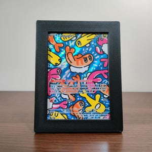 May include: A framed Tatsugiri Pokémon trading card. The card features a colourful illustration with orange, yellow, and pink characters on a blue background. The black frame has a textured appearance and is displayed on a wooden surface.
