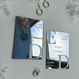 Pode incluir: Conjunto de convites de casamento com um convite azul e espelhado. Os nomes "Emma e Daniel" são exibidos. O conjunto inclui brincos e um anel. Flores secas e um selo de cera adornam o convite.