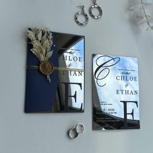 Puede incluir: Elegante conjunto de invitaciones de boda. Tarjetas de acrílico espejado con los nombres "Chloe & Ethan". La tarjeta azul marino está adornada con flores secas y un sello de cera. Pendientes y anillos de plata.
