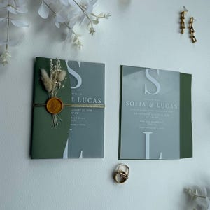 Könnte beinhalten: Zwei Hochzeitseinladungen mit den Namen "Sofia & Lucas" und dem Initial "S". Die Einladungen sind grün und grau mit goldenen Akzenten. Eine Einladung ist mit einem goldenen Wachssiegel versiegelt und mit goldenem Faden gebunden. Die andere Einladung hat eine transparente Folie.