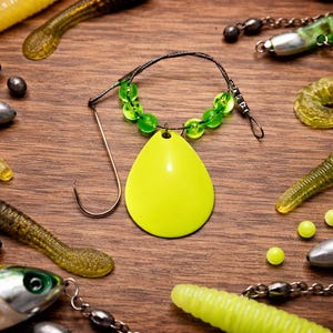 May include: An assortment of fishing lures and accessories displayed on a wooden surface. A bright yellow teardrop-shaped lure with a green beaded necklace and a hook is central. Various soft plastic worms and metal weights are scattered around.