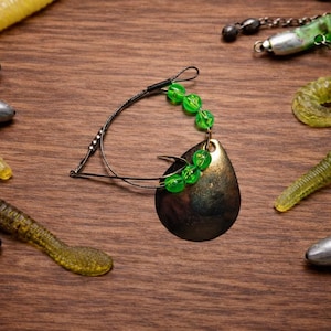 May include: Fishing lures and tackle arranged on a wooden surface. The image features a central spinner bait with a bronze blade and green beads, surrounded by various soft plastic worms and lead weights. The lures are in shades of green and yellow.