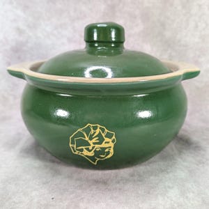 May include: A round, forest green ceramic pot with a matching lid and two side handles. The exterior features a gold-colored line drawing of a stylized face wearing a head covering. The pot has a smooth, glossy finish and a light beige rim.
