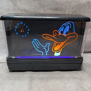 May include: A rectangular black clock featuring a neon-style graphic of a cartoon duck with an orange beak and blue outlines. The clock face has blue numbers and red hands. A horizontal purple neon light tube is positioned at the base of the unit.