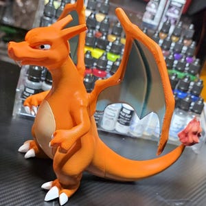 May include: A 3D printed orange dragon-like figure with a cream-colored belly, large wings with teal undersides, white claws, and a flame at the tip of its long tail. The figure stands in a bipedal pose with a sharp, focused facial expression.