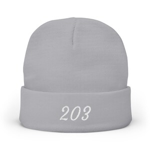May include: A light gray beanie with a folded brim. The number "203" is embroidered in white script on the front of the brim. The beanie is made of a soft, knitted material.