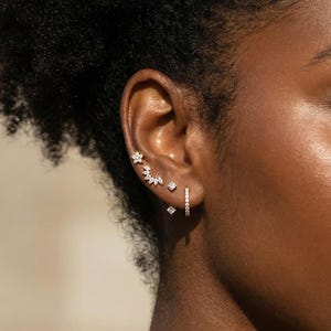 Puede incluir: Primer plano de una oreja adornada con varias joyas. La oreja presenta un conjunto de pequeños y brillantes pendientes, incluyendo diseños florales y geométricos, y un pequeño pendiente de aro. Las joyas se destacan sobre la piel y el cabello de la persona.