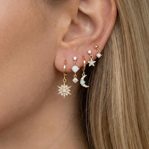 Könnte beinhalten: Eine Auswahl an goldfarbenen Ohrringen mit Himmelsmotiven. Die Ohrringe umfassen einen Sonnenaufgang, einen Mond, einen Stern und geometrische Formen, alle mit kleinen, funkelnden Akzenten verziert. Die Ohrringe werden an einem Ohr gezeigt, was ihr zartes und stilvolles Aussehen unterstreicht.