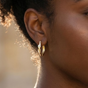 May include: Close-up of a gold hoop earring with a twisted design and small, sparkling embellishments. The earring is worn on a person's ear, showcasing its intricate details and warm, golden hue. The earring's design adds a touch of elegance.