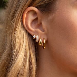 May include: A close-up of a person's ear adorned with multiple gold earrings. The set includes a small square-cut stud, a hoop with small stones, and two different sized gold hoops. The earrings are shiny and reflect light.