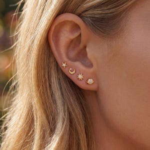 May include: A close-up of an ear adorned with four gold-colored stud earrings. The earrings feature celestial designs: a star, a crescent moon, a sun, and a planet. Each earring is embellished with small, sparkling stones.