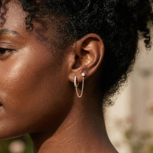 以下が含まれることがあります： 2つのジュエリーで飾られた耳のクローズアップ。1つは、小さな透明な石が付いた小さな金のフープイヤリングです。もう1つは、繊細なゴールドチェーンでフープに接続された小さな丸いスタッドイヤリングです。