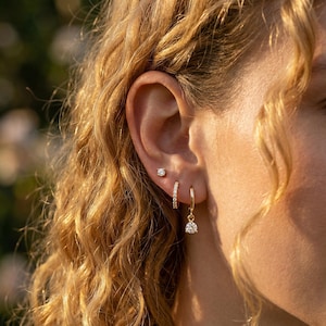 Puede incluir: Primer plano de una oreja adornada con tres pendientes. El pendiente superior es un pequeño y redondo botón. El pendiente del medio es un pequeño aro con piedras incrustadas. El pendiente inferior es un aro dorado con una piedra clara redonda colgante.