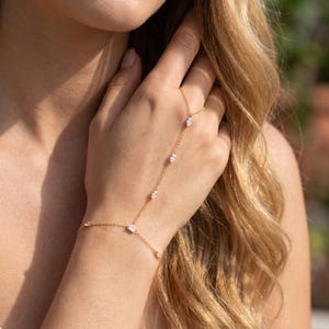 Puede incluir: Una pulsera de cadena de mano dorada con múltiples piedras preciosas claras en forma ovalada. La pulsera se conecta a un anillo y se usa en la mano. La joyería se muestra en la mano de una persona con cabello largo y rubio en el fondo.