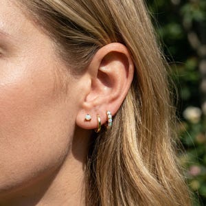 Peut inclure: Gros plan sur une oreille ornée de trois boucles d'oreilles dorées. Une petite puce avec une opale blanche, un simple anneau et un anneau avec plusieurs opales blanches. Les boucles d'oreilles se détachent sur un fond flou.